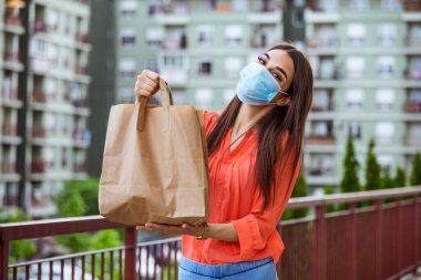 Contactless delivery of goods during Covid 19, coronavirus pandemic. The food delivery courier is holding a large paper bag in her hands. Woman with medical mask delivering food.