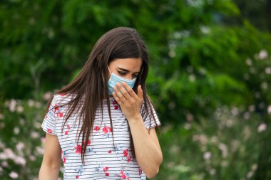 Portrait of young woman wearing a protective mask sneezes on green nature background with flowers in protective medical mask, coronavirus pandemic