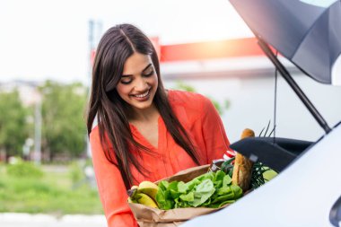 Woman putting her shooping bags into car at shopping mall parking lot. She has done some light shopping,mainly food. Young happy beautiful woman shifts the purchase from shopping cart in the trunk of a car on the parking
