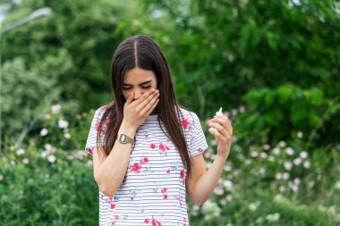 Young pretty woman sneezing in front of blooming tree. Spring allergy concept. Woman applies nasal spray. Pollen allergy symptoms