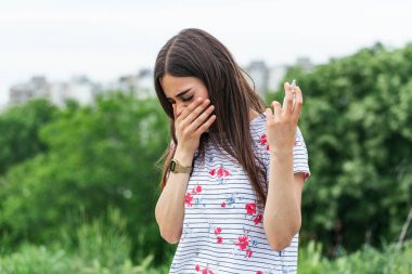 Concept of spring illness. Young female holding napkins and spray for her allergy or fever, cold and running nose in blooming garden. Irritated woman with seasonal allergy
