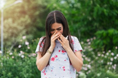 Young pretty woman sneezing in front of blooming tree. Spring allergy concept. Seasonal allergies and health problems. Sinus ache causing very paintful headache. Flu cold symptom. Sinus pain
