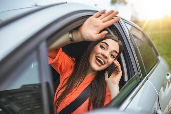 Young woman talking on the phone in the car and waving. Close up portrait of young business woman sitting in the car and laughing while talking on mobile phone