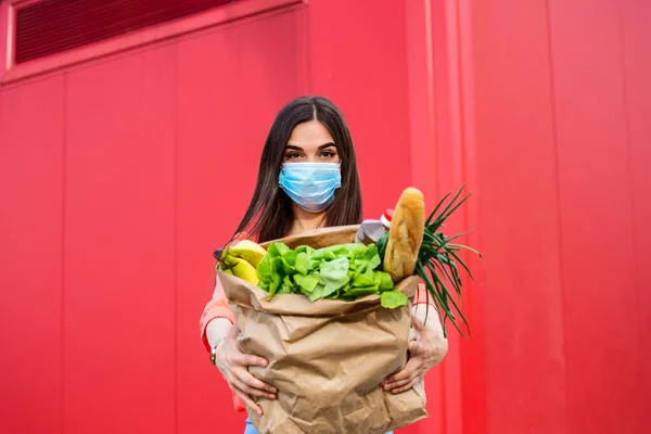 Young woman in medical mask holds an eco paper bag with food, fruits and vegetables, pepper, baguette, lettuce, safe online smart delivery, coronovirus, quarantine, pandemic, stay home concept