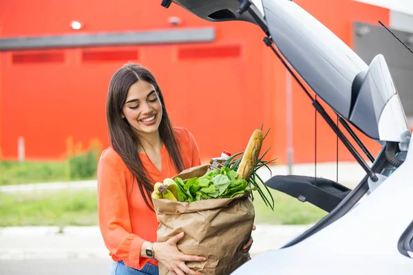 Caucasian brunette going holding paper bags with food products. Young woman putting package with groceries and vegetables into car trunk. Attractive caucasian female shopping in mall or grocery store