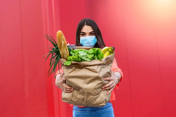 Young woman in medical mask holds an eco paper bag with food, fruits and vegetables, pepper, baguette, lettuce, safe online smart delivery, coronovirus, quarantine, pandemic, stay home concept