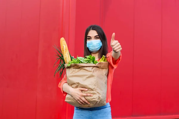 Pretty girl with medical mask holding bag with groceries and looking at camera with thumb up. Woman holding heavy bag with groceries. Groceries shopping during Covid 19, coronavirus pandemic