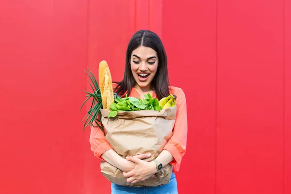 Beautiful woman holding grocery shopping bags on red background . Happy pretty girl holding bag with groceries over red background
