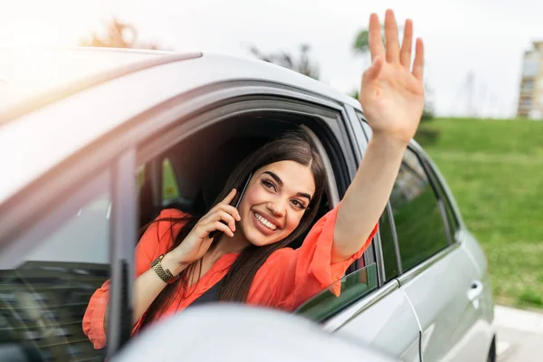 Young woman talking on the phone in the car and waving. Close up portrait of young business woman sitting in the car and laughing while talking on mobile phone