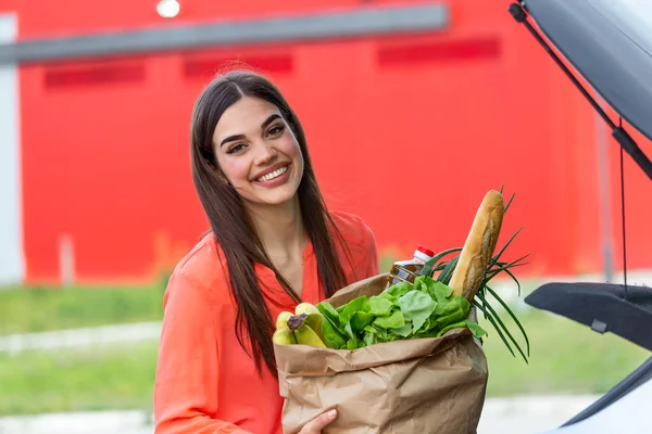 Caucasian brunette going holding paper bags with food products. Young woman putting package with groceries and vegetables into car trunk. Attractive caucasian female shopping in mall or grocery store