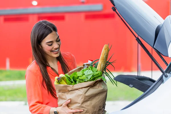 Caucasian brunette going holding paper bags with food products. Young woman putting package with groceries and vegetables into car trunk. Attractive caucasian female shopping in mall or grocery store