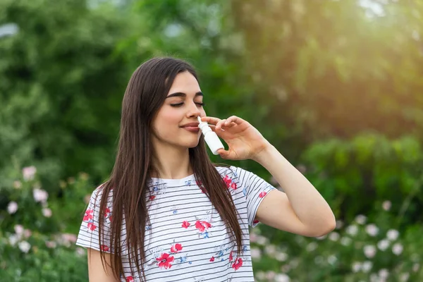 Pollen allergy concept. Young woman using nose spray for her pollen and grass allergies. Flowering trees in background. Spring Seasonal allergies and health problems.