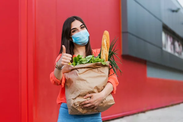 Pretty girl with medical mask holding bag with groceries and looking at camera with thumb up. Woman holding heavy bag with groceries. Groceries shopping during Covid 19, coronavirus pandemic