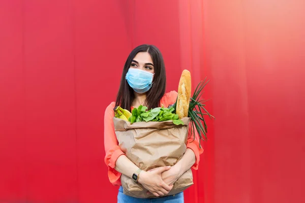 Young woman in medical mask holds a paper bag with food, fruits and vegetables, pepper, baguette, lettuce over grey background, home delivery, coronovirus, quarantine, stay home concept, copy space