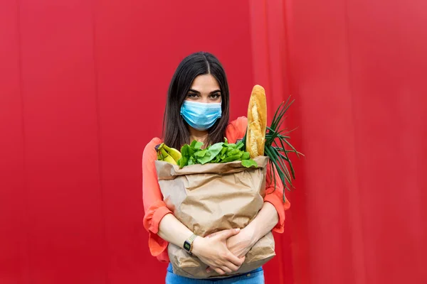Girl courier volunteer in a medical mask holds a paper bag with products, vegetables, herbs isolated over orange background, quarantine, coronavirus, safe food delivery