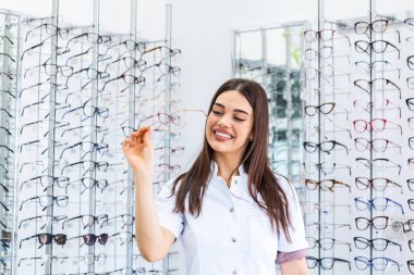 Attractive young female doctor in ophthalmology clinic. Doctor ophthalmologist is standing near shelves holding eyeglasses. clipart