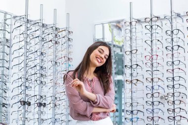 Health care, eyesight and vision concept - happy woman choosing glasses at optics store. Young beautiful girl ar optic store trying on new glasses