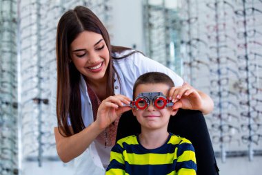 Attractive ophthalmologist examining youg boy with optometrist trial frame. Kid patient to check vision in ophthalmological clinic. clipart