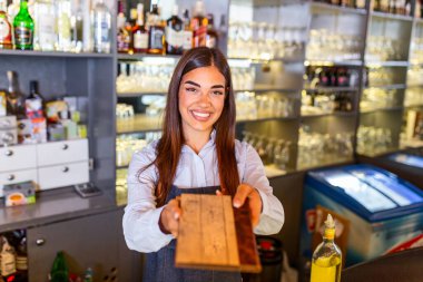 Happy beautiful smiling waitress wearing apron holding a folder menu in a pub, looking at camera, standing in cozy coffeehouse, good service