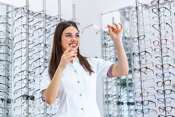 Attractive young female doctor in ophthalmology clinic. Doctor ophthalmologist is standing near shelves with different eyeglasses. Young Doctor in optic store testing glasses