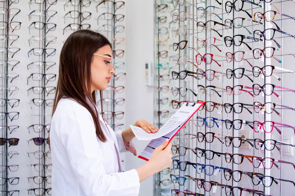 Привлекательная женщина-офтальмолог записывает данные в буфер обмена, работает в оптическом магазине. Здравоохранение и медицина
