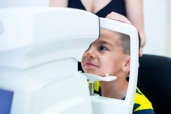 Female oculist using machine for checking eye sight in clinic. Little boy looking at equipment and doctor testing eye pupil in optical store. Concept of eye care and health