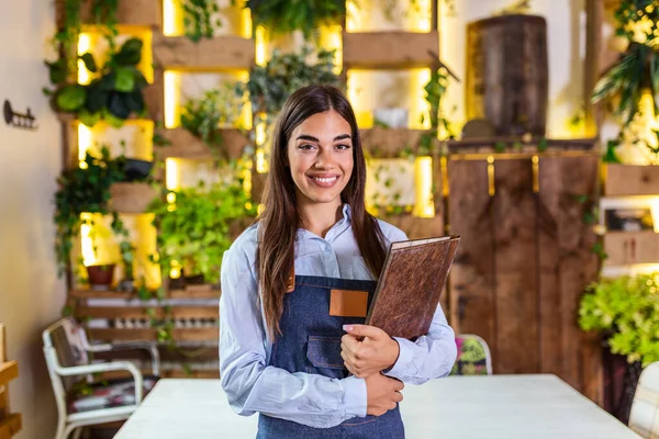 Happy beautiful smiling waitress wearing apron holding a folder menu in a restaurant, looking at camera, standing in cozy coffeehouse, good service