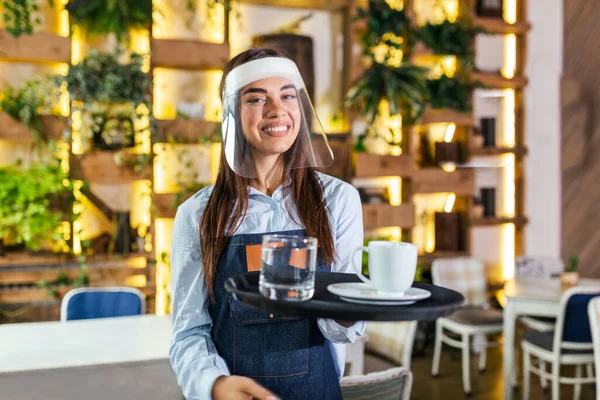 Female waitress wearing face shield, visor serves the coffee in restaurant during coronavirus pandemic. Holding tray with cup of coffee, working in cafeteria and serves the table