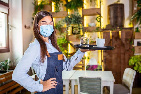 Happy waitress holding tray with cup of coffee, working in cafeteria and serves the table. Young woman wearing protective face mask during coronavirus pandemic