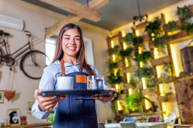 Cheerful young waitress holding tray, smiling happy, looking at camera. Happy waitress wearing apron, holding tray, working in cafeteria or restaurant