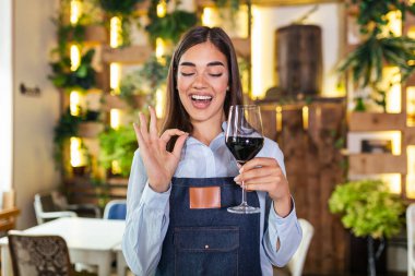 Joyful elegant waitress holding glass of red wine showing delicious sign , standing in restaurant. Sommelier tasting wine in restaurant.