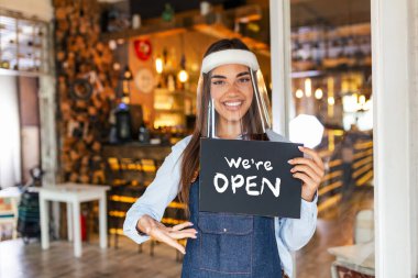 Small business owner smiling while holding the sign for the reopening of the place after the quarantine due to covid-19. Woman with face shield holding sign we are open, support local business.