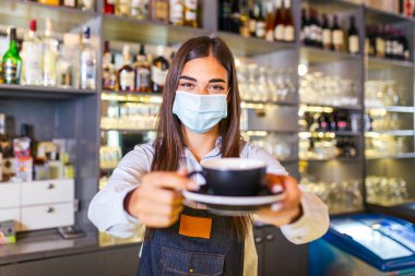 Beautiful female barista is holding a cup with hot coffee, looking at camera and wearing protective face mask while standing near the bar counter in cafe