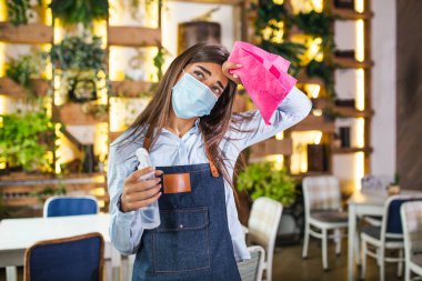 Portrait of attractive female waitress wearing face mask holding a bottle with sanitizer and a rag in a restaurant. New normal with coronavirus outbreak hygiene restaurant concept.