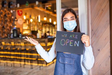 Happy female waitress with protective face mask holding open sign while standing at cafe or restaurant doorway, open again after lock down due to outbreak of coronavirus covid-19