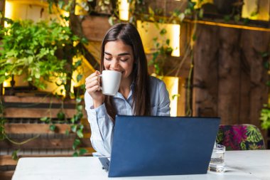 Young business woman in sitting at table in cafe and drinking coffee. On table is laptop and smartphone Freelancer working in coffee shop. Student learning online.