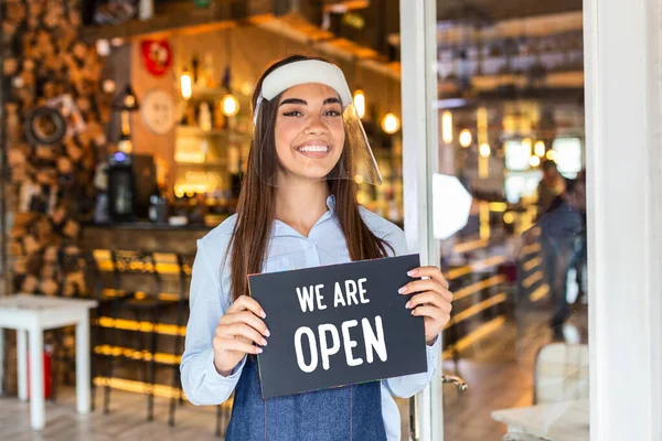 Small business owner smiling while holding the sign for the reopening of the place after the quarantine due to covid-19. Woman with face shield holding sign we are open, support local business.