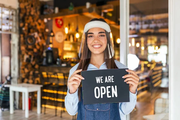 Small business owner smiling while holding the sign for the reopening of the place after the quarantine due to covid-19. Woman with face shield holding sign we are open, support local business.
