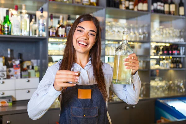 Beautiful female bartender is holding a shot glass with alcohol drink and a bottle in other hand, looking at camera and smiling while standing near the bar counter in cafe