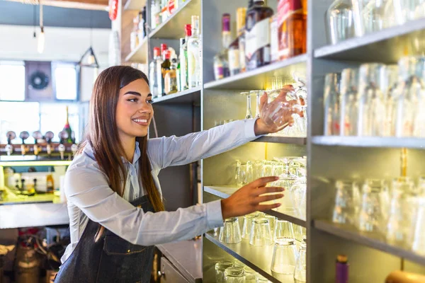 Bartender puting clean glass on the shelf. Young woman behind bar taking a clean glass to pour a drink. Bartender in the bar or restaurant packing glasses