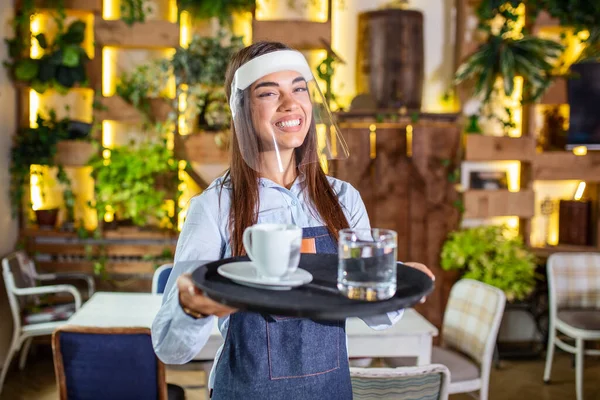 Female waitress wearing face shield, visor serves the coffee in restaurant during coronavirus pandemic . Holding tray with cup of coffee, working in cafeteria and serves the table