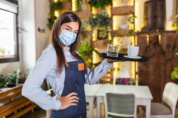 Female waitress in a medical protective mask serves the coffee in restaurant durin coronavirus pandemic representing new normal concept