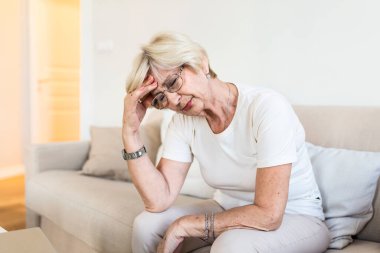 Mature woman sitting on a white sofa in a home touching her head with her hand while having a headache pain and feeling unwell
