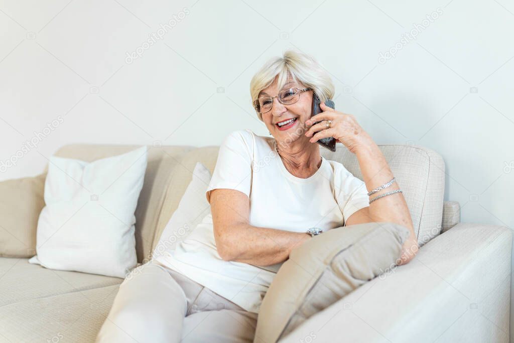 Beautiful Caucasian elderly female having phone conversation with her old friend using electronic device, discussing latest news with cute smile on her face, sitting at home