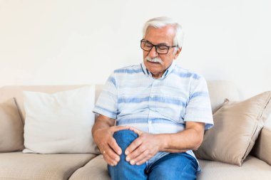 Elderly man sitting on a sofa at home and touching his knee by the pain.