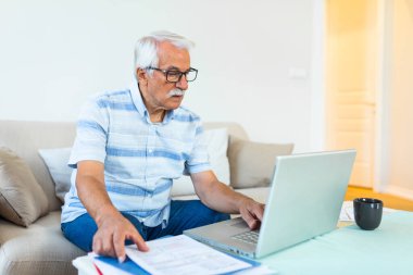 Odaklanmış gri saçlı yaşlı adam iç harcamaları hesaplayan banka bildirimleri okuma kanepede oturup, konsantre modern olgun erkek mali evrak düşünün, dizüstü bilgisayar online faturaları ödemek
