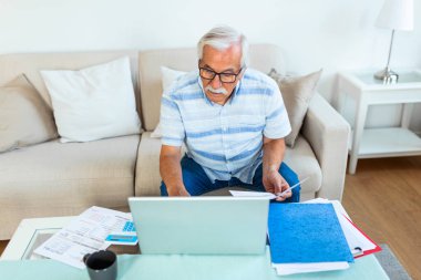 Elderly man sitting on couch at home planning budget check bills cheques, computer, documents and calculator on coffee table, close up cropped concept image, old man manage family expenses concept