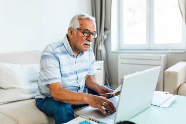 Old retiree man sit on couch holding receipt paper calculates on calculator monthly expenses, elderly male managing budget, planning finances, analyzes charges, check utility bills concept