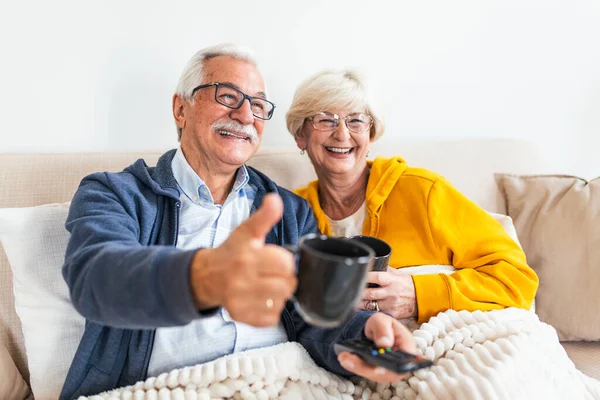 Senior couple sitting on the sofa,covered with blanket. feeling cozy, watching television. Elderly man showing thumb up