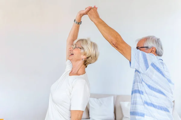 Carefree happy active old senior couple dancing jumping laughing in living room, cheerful retired elder husband holding hand of mature middle aged wife enjoy fun leisure retirement lifestyle at home